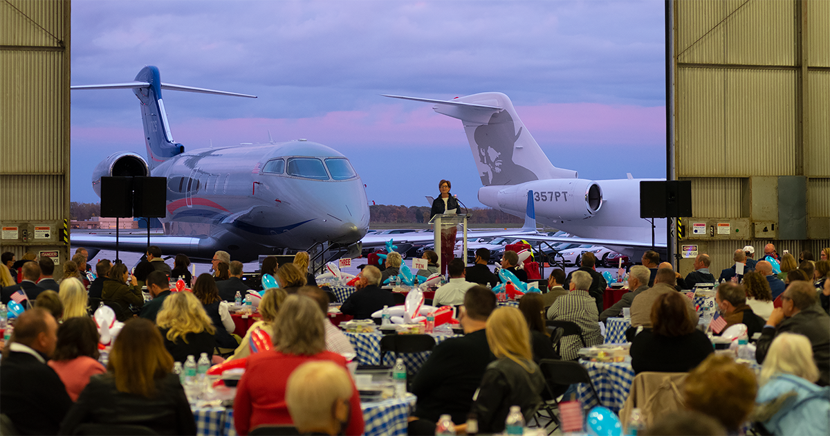 Two airplanes sit in the background while a person delivers a speech during the GFW Inc. Annual Meeting at the Fort Wayne Aero Center