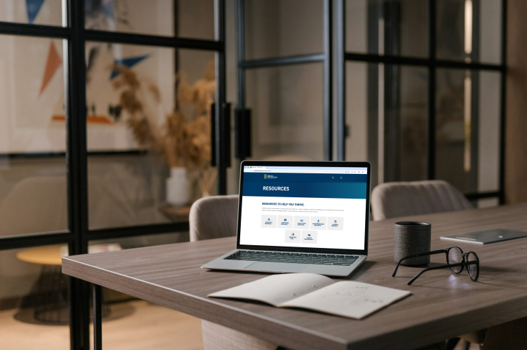 Laptop computer sitting on a table with the Greater Fort Wayne Inc. Resources webpage on the screen. a mug, glasses, and a book are sitting around it.