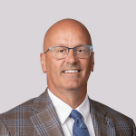 Middle-aged, caucasian male. Bald with glasses, wearing a tan patterned suit and blue tie.